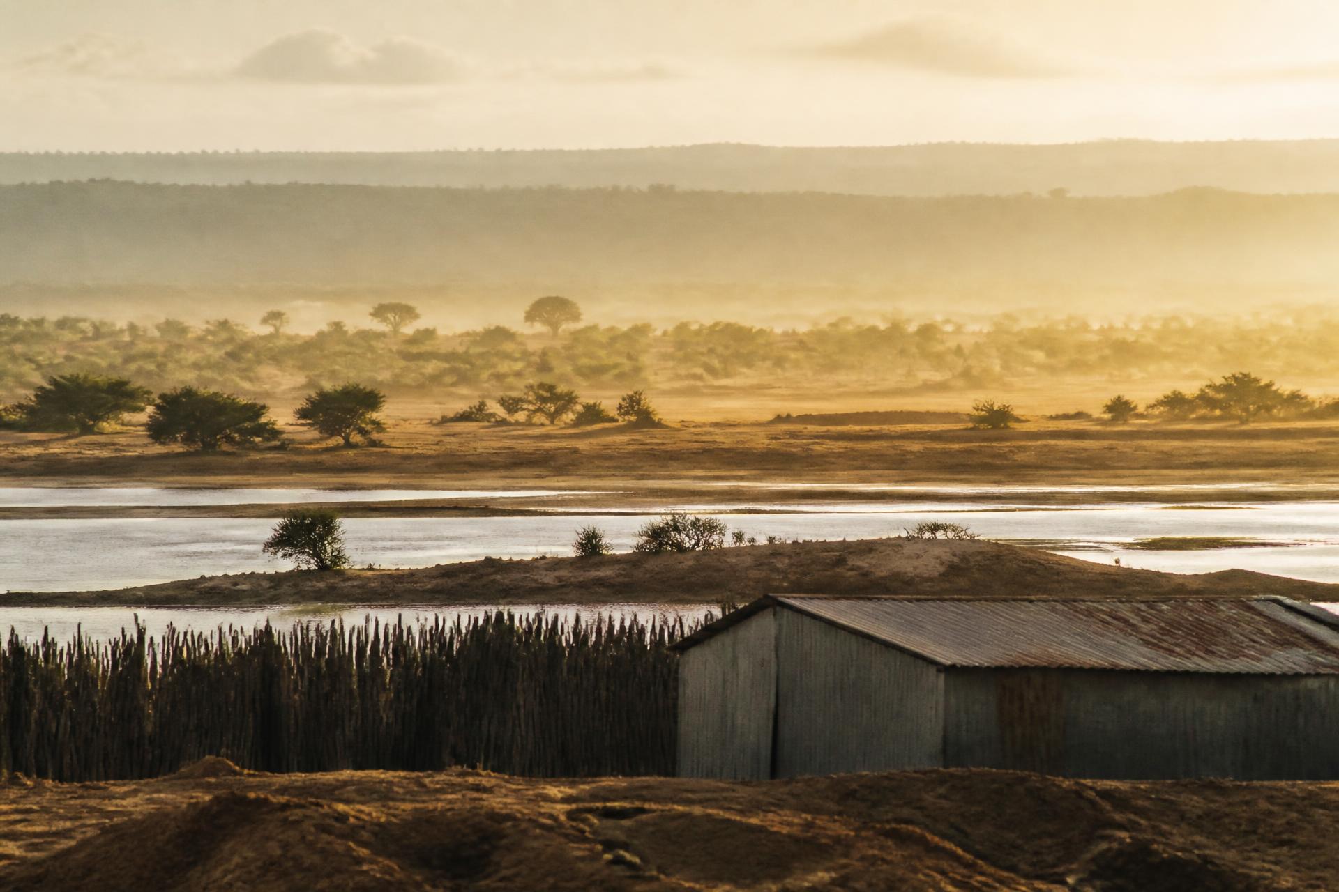 Low sun over a Tanzanian floodplain, with a timber fence and a metal-roofed store beside still water.